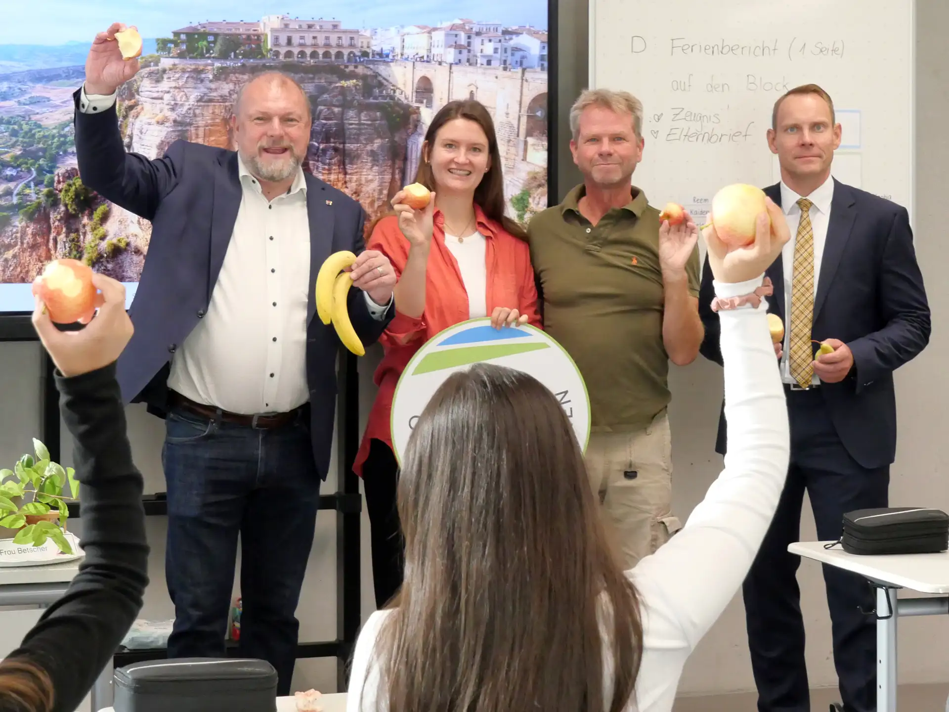 Person verteilt Äpfel, Birnen und Bananen an Schülerinnen und Schüler vor einem Schulgebäude.