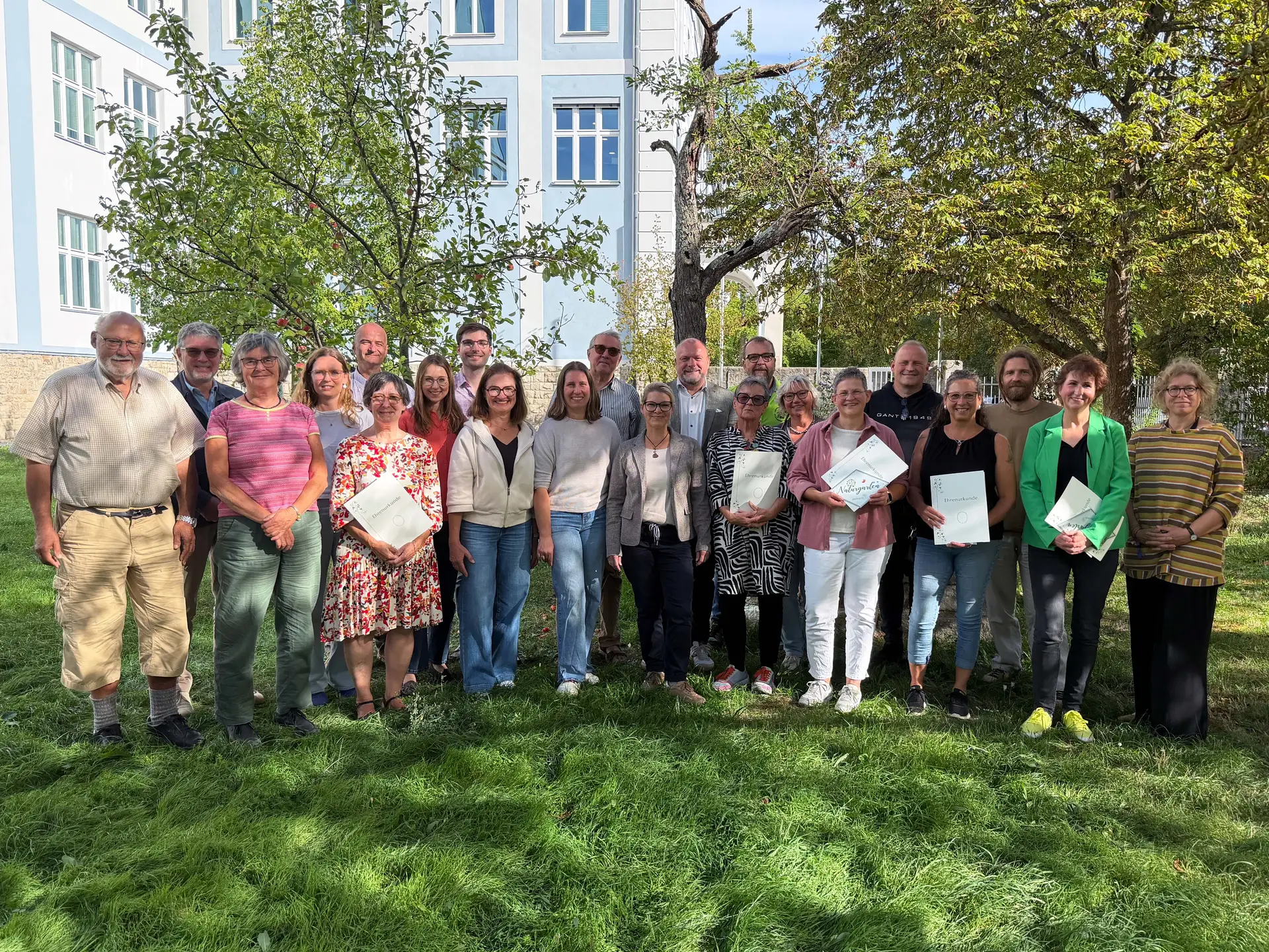 Eine größere Gruppe steht auf einer Wiese vor einem Gebäude; mehrere Personen halten Urkunden zur Naturgarten-Auszeichnung.